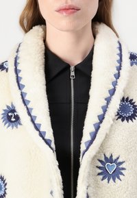 Luddig elfenbensfärgad jacka med blå broderade blommönster, kontrasterande zigzagkant och ett svart dragkedjeinnerlager.