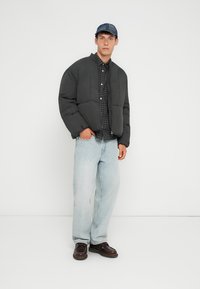 Sort polstret jakke, rundede skuldre, gråternet skjorte under, lyseblå jeans og mørkebrun sko. Modellen står foran en hvid baggrund.