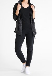 Ensemble de sport noir comprenant un sweat à capuche zippé à texture rayée, un haut sans manches et un pantalon fuselé, associé à des baskets blanches.