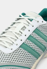 Chaussure de sport gris clair avec une tige en mesh, des accents bleu sarcelle, des lacets blancs, et trois bandes caractéristiques sur le côté.