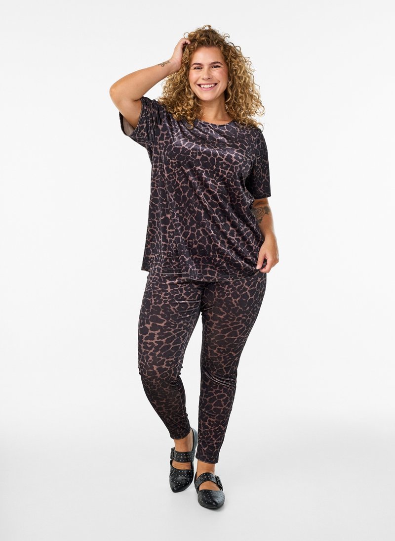 Smilende kvinde med krøllet hår står iført matchende mørk leopardprint top og leggings med sorte flade sko med nitter på hvid baggrund.
