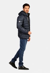 Doudoune marine avec capuche, présentant des coutures horizontales, des poches latérales et un écusson logo sur la manche gauche. Portée avec un jean bleu et des bottes noires.