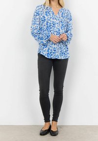 Niebieska bluzka w kwiaty z dekoltem w kształcie litery V, długimi rękawami i marszczonymi mankietami, zestawiona z wąskimi czarnymi dżinsami i czarnymi płaskimi butami.