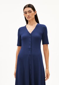 Robe en maille bleu marine avec des manches courtes, un décolleté en V et des détails de bouton à l'avant. Corsage ajusté qui s'évase vers une jupette fluide. Tissu texturé et finition lisse.