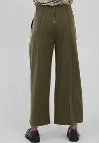 Pantalons larges vert olive avec une texture lisse, dotés d'une fermeture éclair dans le dos et de poches latérales, associés à des chaussures noires.