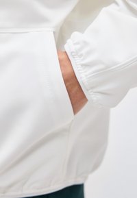 Το χέρι ενός ατόμου είναι μερικώς μέσα στην πλαϊνή τσέπη ενός λευκού μπουφάν με ελαστικές μανσέτες και τελείωμα.