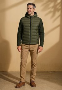 Olijf groene gevoerde hoodie met een doorgestikt voorpaneel, zwarte mouwen, ritsluiting, gecombineerd met tan chino's en bruine leren schoenen, staande binnen.