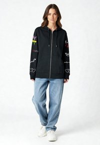Čierna mikina s kapucňou na zips s farebnou výšivkou na rukávoch, zdobená kvetmi a nápismi. Kombinovaná s voľnými svetlomodrými džínsami a bielymi teniskami.