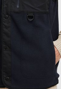 Chaleco de forro polar navy con acentos en negro, que presenta un gran bolsillo frontal, botones de presión y un detalle en forma de lazo. Textura suave con un acabado detallado.