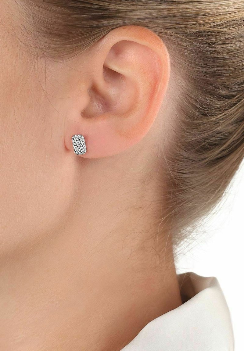 Silverfärgad ploppörhänge med en texturerad rektangulär design och små glittrande detaljer. Bärs på örat mot ljus hy.