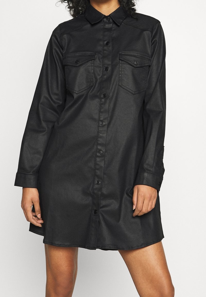 Femme portant une robe-chemise noire à manches longues boutonnée avec des poches poitrine, debout devant un fond uni.