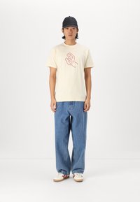 Romantische katoenen t-shirt in crème met een rood handgrafiek, gecombineerd met losse blauwe denim jeans en witte sneakers met rode strepen. Zwarte pet gedragen.