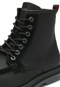 Stivale alla caviglia nero con tomaia in tessuto testurizzato, dettagli in suede, occhielli in metallo, lacci piatti e un tab di richiamo rosso a contrasto sul tallone.