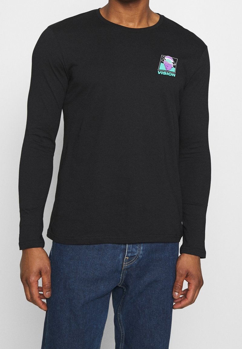 Homme portant une chemise noire à manches longues avec un petit graphique violet et bleu sarcelle représentant une tête et le mot « VISION » sur le haut du côté gauche de la poitrine.