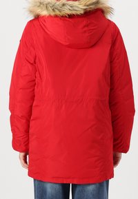 Rød dunjakke med aftagelig hætte foret med pels, med indsnævret talje og lange ærmer for ekstra varme. Tekstureret stof, midt-lang.