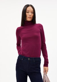 Burgunderfarbener Rollkragenpullover mit vertikalen Streifen, aus weichem Stoff; kombiniert mit dunklen Jeans mit Fronttaschen und Knopfverschluss.