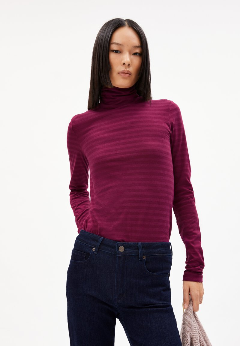Burgunderfarbener Rollkragenpullover mit vertikalen Streifen, aus weichem Stoff; kombiniert mit dunklen Jeans mit Fronttaschen und Knopfverschluss.