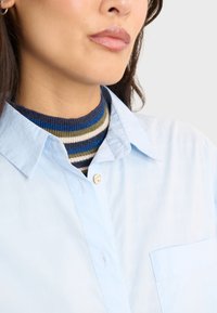 Camicia azzurra chiara con taschino sul petto, indossata sopra un maglione a righe multicolori nei toni blu navy, verde e blu.