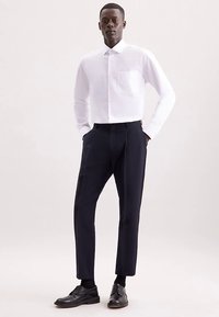 Witte overhemd met lange mouwen en een borstzak, gecombineerd met donkerblauwe broek. Model draagt zwarte nette schoenen, gefotografeerd tegen een neutrale achtergrond.