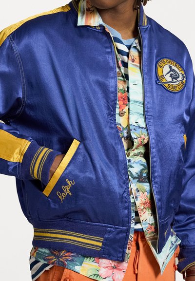 Blau Bomberjacke mit gelben Akzenten, gesticktem Logo und gestreiften Bündchen. Getragen über einem blumigen Hemd mit lebendigen Farben.