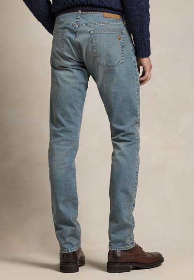 Zadní pohled na osobu v světlých úzkých džínách, tmavě modrém svetr s copánkovým vzorem a tmavě hnědých kožených botách na neutrálním pozadí.