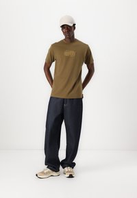 Oliven grønn bomulls T-skjorte med et grafisk logo, sammen med løse mørkeblå denimjeans og nøytrale joggesko. Lys caps på hodet.
