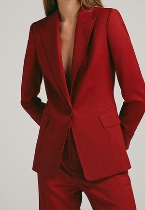 Nainen pukeutuneena räätälöityyn, napitettuun punaiseen jakkuun ja siihen sopiviin housuihin, kädet selän takana, taustalla yksivärinen tausta.