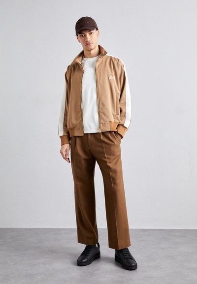 Giacca marrone con zip e strisce laterali bianche, indossata sopra una camicia bianca, abbinata a pantaloni marroni e sneaker nere. Berretto piatto sulla testa.
