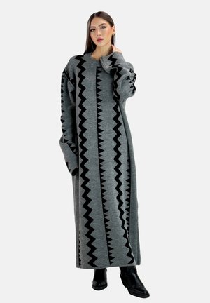 Frau mit langen dunklen Haaren, die ein langes graues Strickkleid mit schwarzen Zickzackmustern und schwarze Stiefeletten trägt, posiert mit einer Hand am Hals.