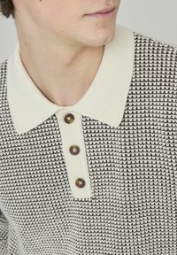 Polo en tricot avec un motif texturé crème et marron, col côtelé et trois boutons marron sur le plastron.