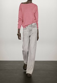 Jasnoróżowy sweter z odkrytymi ramionami w połączeniu z luźnymi, jasnoszarymi, wysokim stanem jeansami. Modelka ma na sobie płaskie sandały i kilka bransoletek.