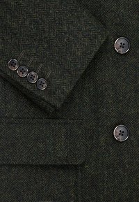 Ciemnozielona marynarka w jodełkę z wełny, z czterema czarnymi guzikami przy mankietach oraz kieszenią. Teksturowany materiał o plecionej strukturze.