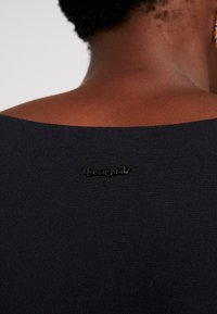 Mustat kankaatoppi, jossa on sileä rakenne ja leveä pääntie; pieni musta "Desigual"-logotunnus takana.