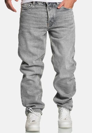 Homme portant un jean gris ample et des baskets blanches, debout avec une main dans la poche sur fond blanc.