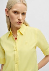 Gelbes kurzärmliges Hemd mit Knopfleiste und glänzender Oberfläche. Verfügt über ein gesticktes "HUGO"-Logo auf der Brust und ein klassisches Kragen Design.