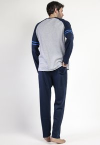 Hombre de pie descalzo, vistiendo una camiseta de manga larga azul marino y gris, y pantalones azul marino, con una mano en el bolsillo, de espaldas frente a un fondo blanco.