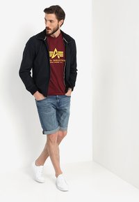 Marine let jakke, rød t-shirt med gul logo, denim shorts og hvide sneakers. Modellen poserede mod en ensfarvet baggrund.