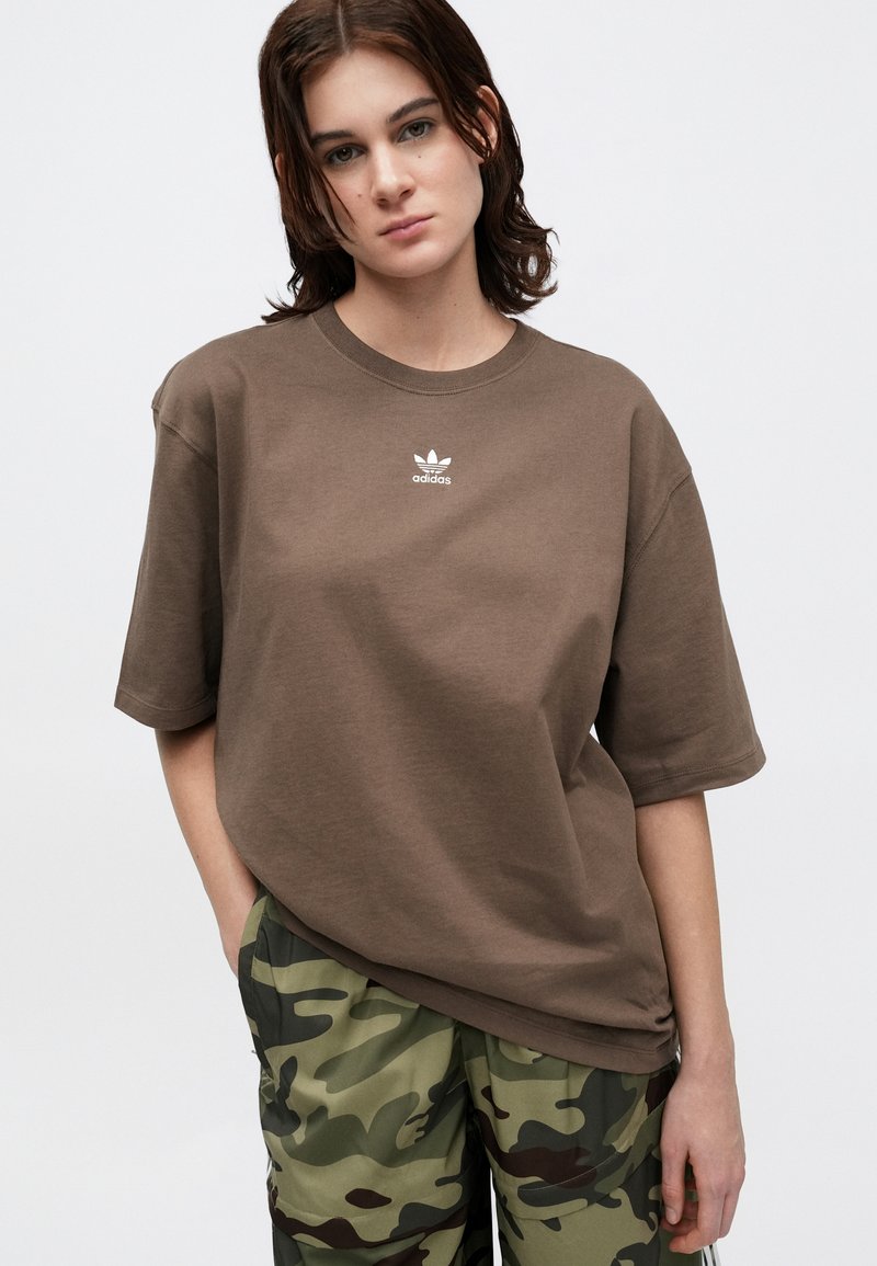 Braunes Baumwoll-T-Shirt mit kurzen Ärmeln, Rundhalsausschnitt und weißem Logo auf der Brust. Kombiniert mit Camouflage-Hosen. Glatte Textur.