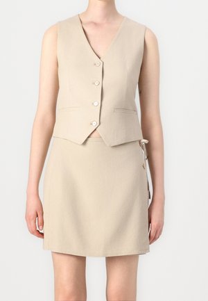 Ærmeløs beige vest med knapper kombineret med et matchende beige skørt med knapper og bindedetaljer i siden, båret af en stående person.