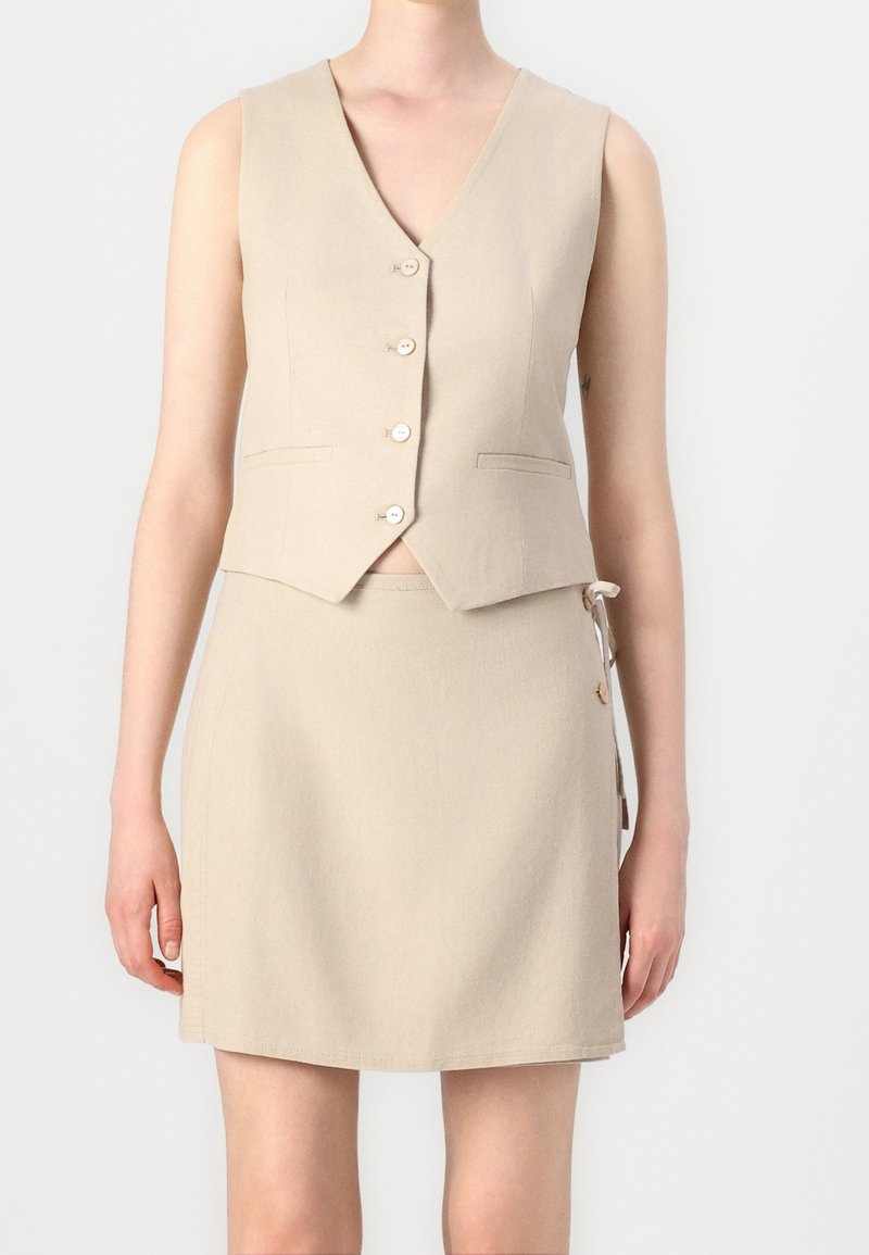 Veste beige sans manches boutonnée, associée à une jupe beige assortie avec des boutons latéraux et des détails de liens, portée par une personne debout.