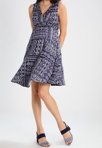 Sleeveless dress with a deep V-neck, featuring a navy and white geometric pattern. Paired with blue and navy block-heeled sandals.
