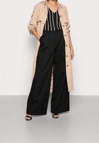 Žena v béžovém trenčku, černých kalhotách s širokými nohavicemi, černobílé pruhované blůze a černých sandálech s otevřenou špičkou, stojí s jednou rukou v kapse.