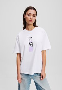 White cotton t-shirt with a relaxed fit, featuring a graphic design in black and purple on the front. Accompanied by light denim pants.