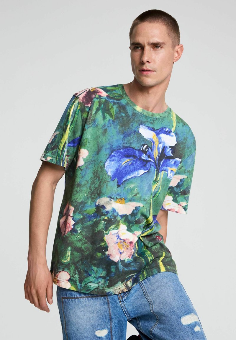 Homme portant un T-shirt vert à motif floral avec de grandes fleurs bleues et roses et un jean bleu déchiré sur un fond uni.