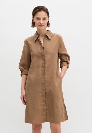Frau trägt ein knielanges braunes Leinenhemd mit hochgekrempelten Ärmeln, steht mit einer Hand in der Tasche vor einem schlichten weißen Hintergrund.
