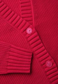 Fuksia neulottu neuletakki, jossa on ribbikuvio, V-kaulus ja kaksi vaaleanpunaista nappia edessä, sekä taskupistoke vasemmalla puolella.