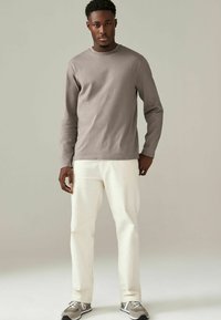 Grijze katoenen shirt met lange mouwen en ronde hals, gecombineerd met crèmekleurige broek. Model draagt grijze sneakers met witte en zwarte accenten.