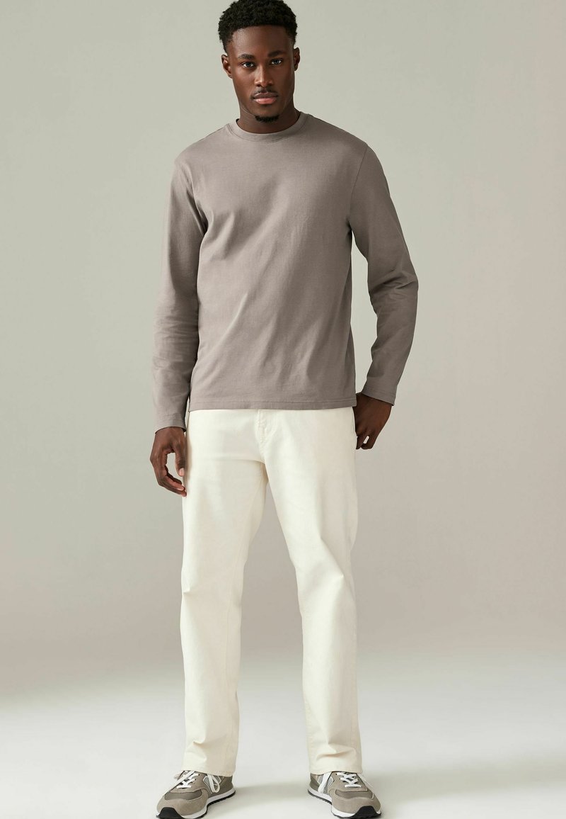 Grijze katoenen shirt met lange mouwen en ronde hals, gecombineerd met crèmekleurige broek. Model draagt grijze sneakers met witte en zwarte accenten.