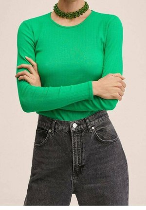 Persoon die een felgroene lange mouw top draagt, hooggetailleerde zwarte jeans en een dikke groene kralenketting, armen gekruist.