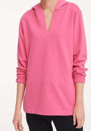 Personne portant un pull rose ample à manches longues avec un col en V profond et une capuche, associé à un pantalon noir.